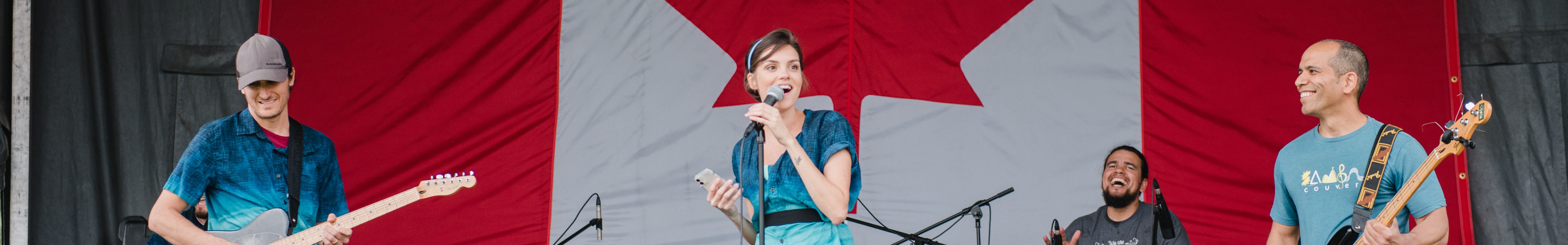 The image depicts a group of people performing on the main stage of a festival. The group includes a drummer, guitarist, bass player, and singer, all holding microphones and smiling. A Canadian flag is visible in the background.
