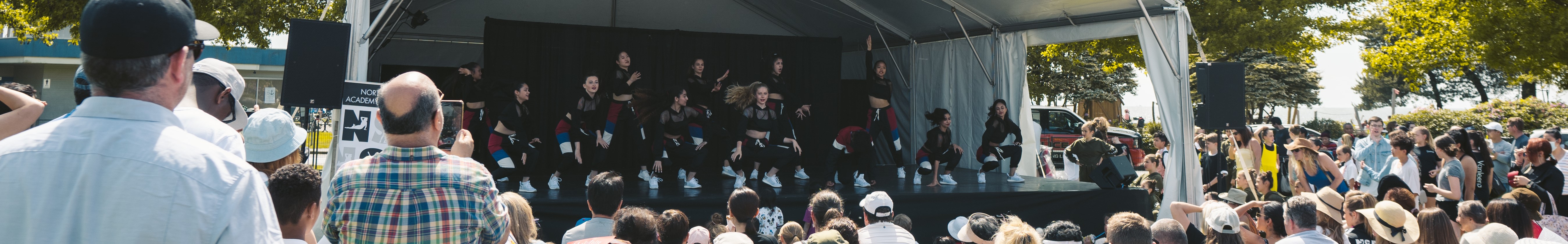 The image is of a group of people on a dance stage at the festival. They are standing and wearing different types of clothing.