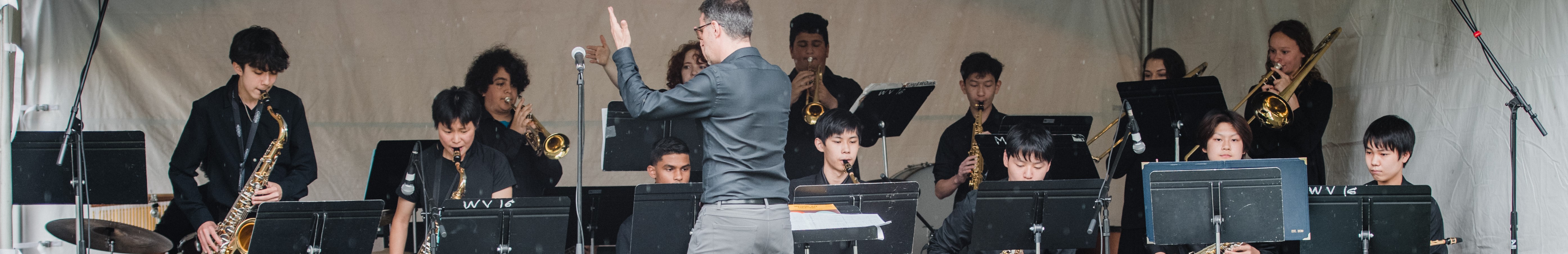 The image shows a group of people playing instruments on the West Vancouver Schools Performance Stage. They are likely a band performing at a concert or musical event.
