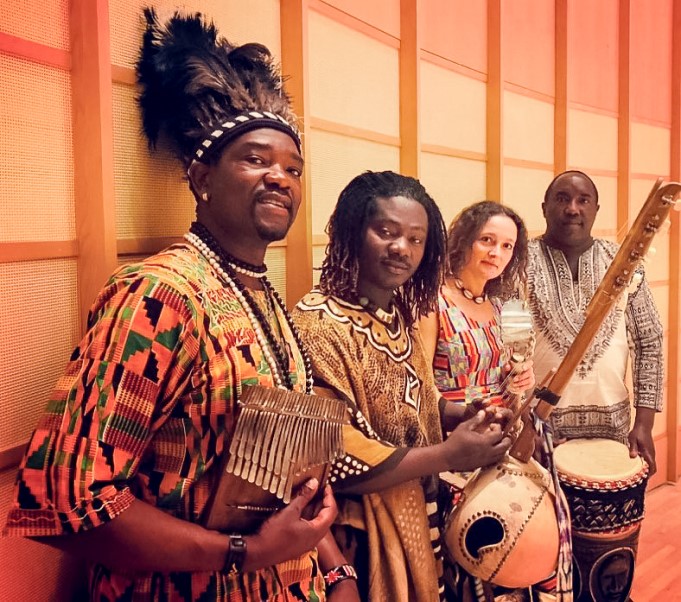 The image shows a group of people holding instruments, including drums and guitars, dressed in African print clothes. They appear to be performing music as part of the Kunda African Culture Music & Dance event. The individuals are standing indoors against a wall, smiling and engaging with the audience.