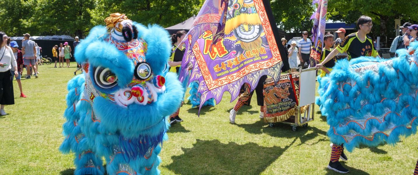 The image features the Vancouver Chinese Lion Dragon Dance Team performing. The team is dressed in traditional clothing and is likely showcasing a dance routine. It is an outdoor event with people watching the performance on grass.