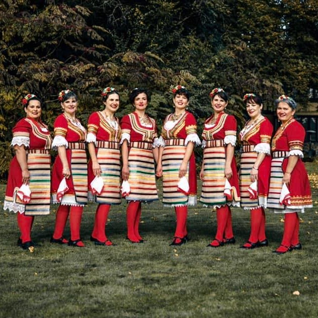 The image features a group of women wearing traditional Bulgarian dress while performing a dance. They are standing in a field with trees in the background. The women are posing and seem to be part of a team or a performance group.