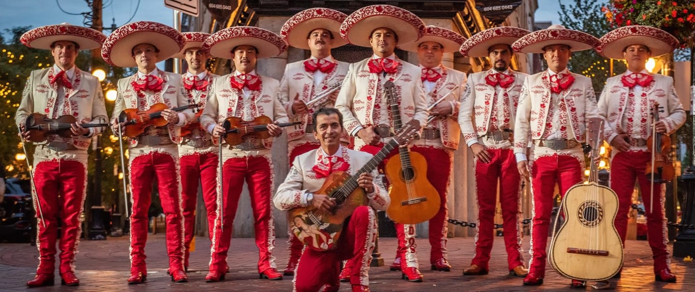 The photo shows a group of people dressed in traditional clothing, likely performing at a festival. The group is called Los Dorados and they play traditional Mexican music with a unique northern twist. The bandleader, vocalist, and guitarist is Alex Alegria.