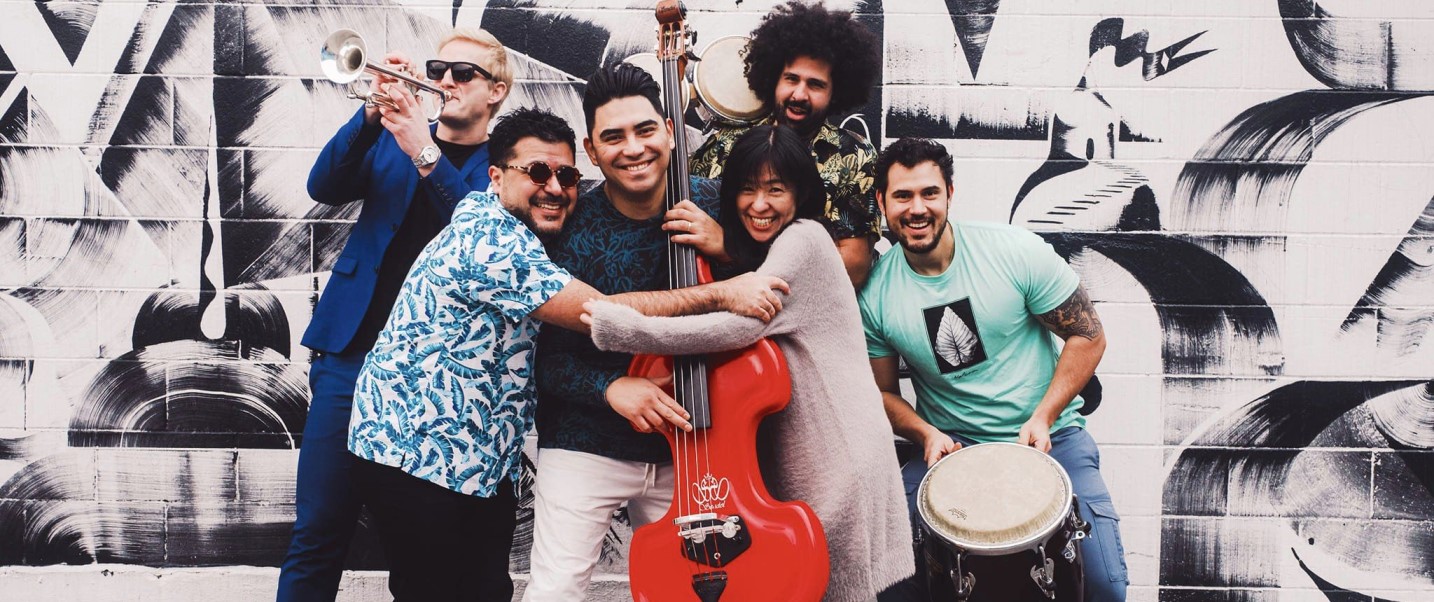 The image depicts a group of people from the Mazacote band posing for a photo outdoors. The group consists of men and women dressed in casual clothing, some holding musical instruments like drums. They are all smiling and standing together for the picture.