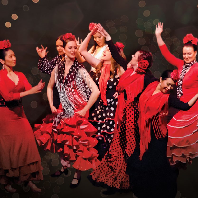 The image shows a group of people dressed in flamenco clothing, likely performing a dance routine. The individuals are wearing vibrant costumes and appear to be smiling. The event is related to Mozaico Flamenco.