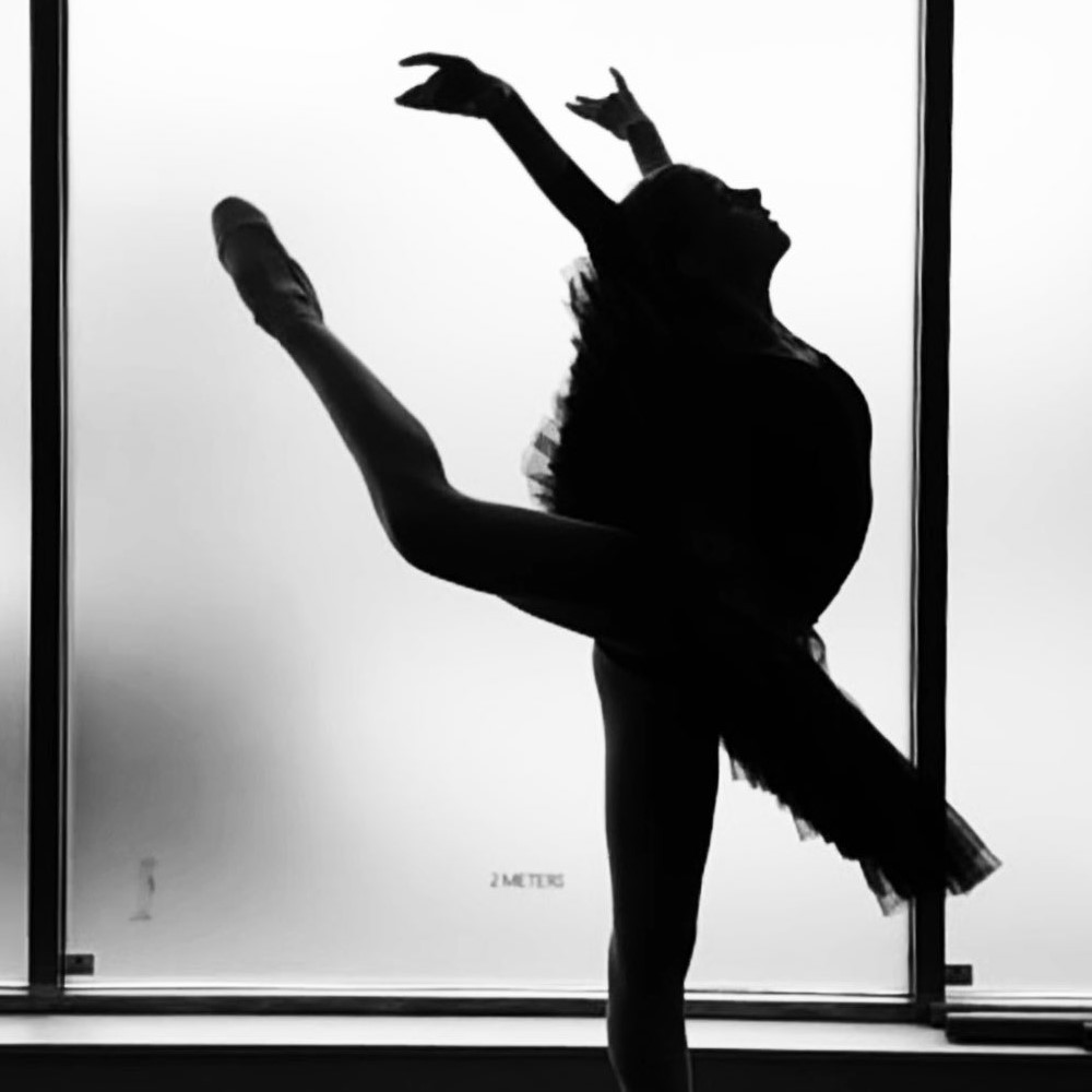 The image shows a woman dancing on a stage, appearing as a silhouette in black and white. The woman is likely a ballerina, showcasing balance and choreography in her performance. 