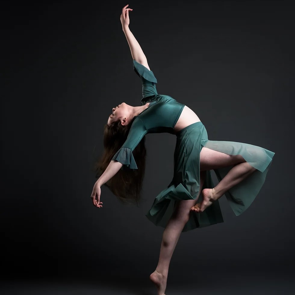 The photo captures a person dancing on a stage at Seymour Dance Academy. The dancer is flexible and wearing a green skirt and blouse, striking an artistic pose. 