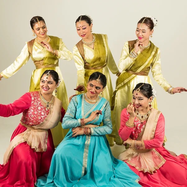 The photo shows a group of women in dresses, likely performing a traditional dance. The women appear to be smiling and engaged in the dance. The setting seems to be indoors, possibly against a decorated wall. The image may be related to a dance performance by the Sri Shakti Academy of Indian Dance.