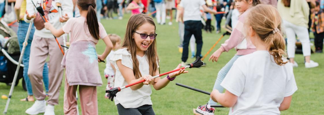 The participants are likely engaged in a circus workshop with the Red Fox Society. The scene includes grass, trees, a ball, and a clear sky. People of different ages and genders are wearing casual clothing and footwear.