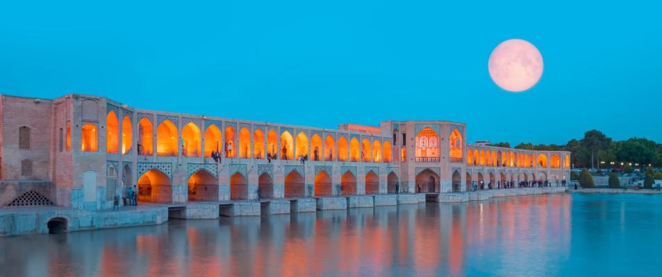 People can be seen resting in the Khaju Bridge in Iran. Key elements include the moon, water, building, sky, and reflection.