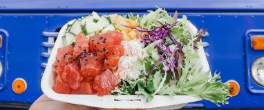 Person holding tray of salmon poke bowl with cucumber, corn, and lettuce in front of blue bus.