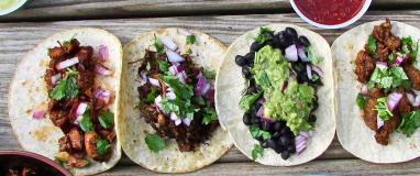 Four tacos on wooden table with onions, cilantro, and guacamole.