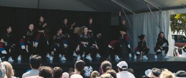 The image is of a group of people on a dance stage at the festival. They are standing and wearing different types of clothing.