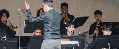 The image shows a group of people playing instruments on the West Vancouver Schools Performance Stage. They are likely a band performing at a concert or musical event.