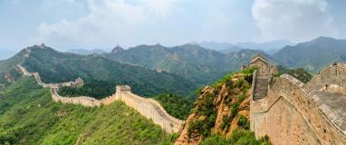 The image is of a stone wall on a hill, part of the Great Wall of China. The setting is outdoors with clouds and sky in the background, surrounded by a landscape with trees and mountains.