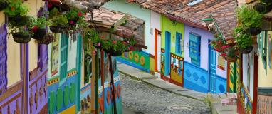 The image shows a street in Guatape, Colombia, with colorful buildings. The buildings are painted in vibrant colors, creating a picturesque scene. The architecture and colors of the buildings give a lively and charming vibe to the street.
