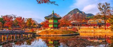 The photo depicts a building, possibly a temple or palace, with a green roof located by a body of water. In the background, there are trees and mountains. This scene is set in Korea.
