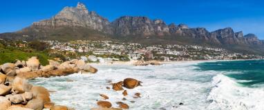 The image shows a rocky beach with a town in the distance. It is a panoramic view of Camps Bay Beach and Table Mountain in Cape Town, South Africa. The landscape includes mountains, the ocean, and rocky coastal landforms.